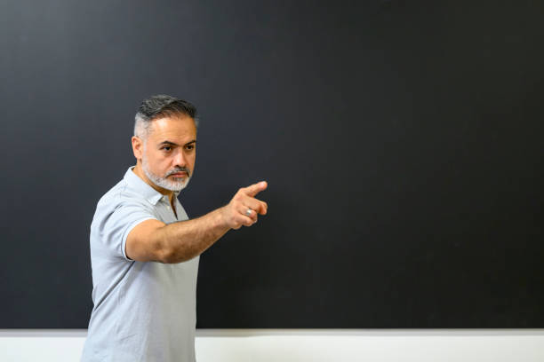 Instructor with blank expression pointing at somebody standing at the blackboard in a classroom