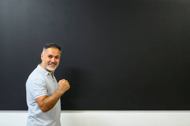Happy smiling instructor making a fist standing at the blackboard in a classroom