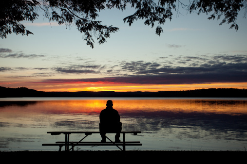 A silhouette of a man sitting on a table and watching a beautiful sunset over a lake. Rear view. A single unrecognizable male watching the sunset over a lake in Ontario, Canada. Sitting on picnic table. Themes include rest, relaxation, single, alone, contemplation, beauty, nature, watching the sunset, people, remote, park, Ontario, lakeside, park, thinking, deep thought, and lifestyle. 