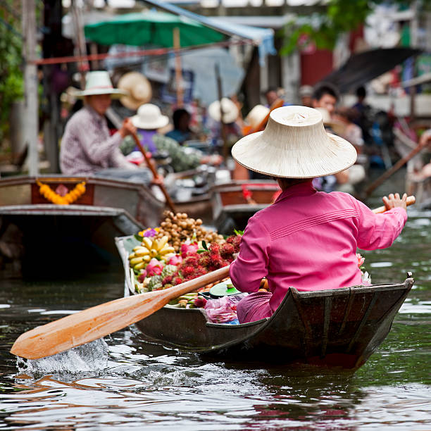 Fruit vendor at a floating market in Thailand A women paddles her floating fruit vendor in a small boat through a waterway in the Damnoen Saduak Floating Market in Thailand.  There are other people selling fruit out of small boats in the foreground. ratchaburi province stock pictures, royalty-free photos & images