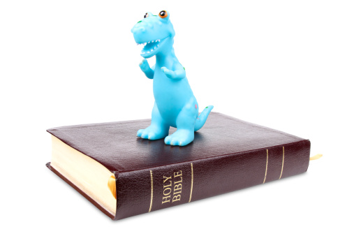 A dinosaur stands triumphantly, on top of the Holy Bible.