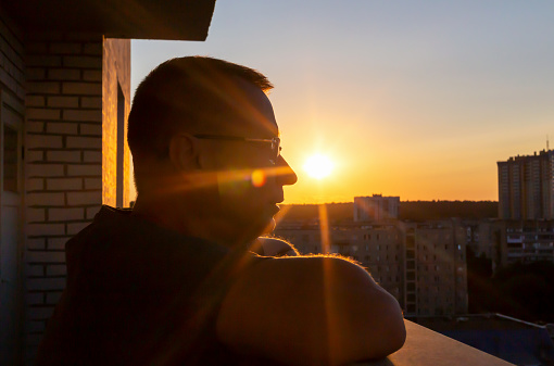 Man in glasses on balcony in rays of setting sun looks forward with hope