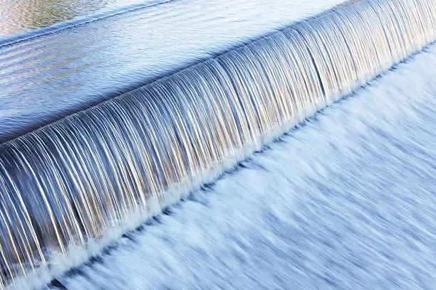 Water flows over a man-made waterfall at the edge of a Lake Placid, New York municipal river dam in a rushing, streaking transition from smooth, rippled calm into frantic, chaotic blur. 