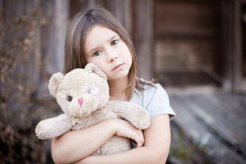 Color photo of a sad, lonely 6-7 year old girl hugging an old, raggedy teddy bear in front of an abandoned house.