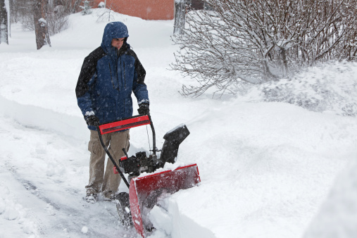 A young man (17 year old teen) clears the driveway with a gasoline-powered snow blower in a suburban neighborhood during a heavy snow blizzard.