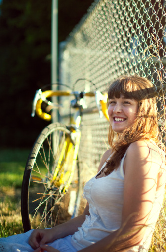 A content young adult girl relaxes against a chain link fence next to her street bike, the warm summer sun shining through from behind.  Vertical with copy space.