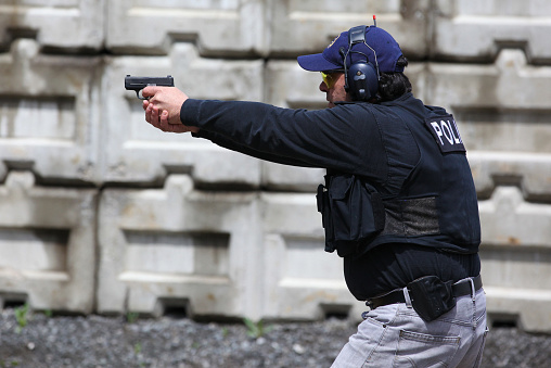 Picture of a man shooting shotgun in a practice field.  The man is wearing a kevlar vest and has earmuffs to protect him from the sound of the handgun. The concrete protective wall fence is in the background. 