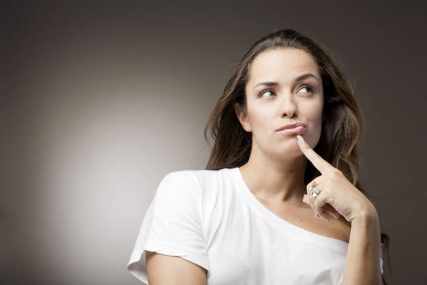 Indoor image of young woman resting chin on hand and sitting behind a glass window reflecting the city street view. She is looking at view outside and at the camera through the glass window with blank expression. One person, headshot and selective focus.