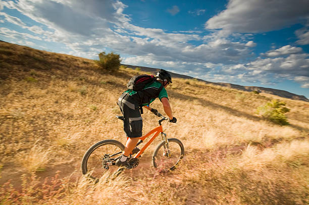 mountain biking! mountain biker speeding past the desert meadow terrain beneath a cloud filled blue sky during sunset turning the landscape a golden yellow.  horizontal composition with copy space. fruita colorado stock pictures, royalty-free photos & images