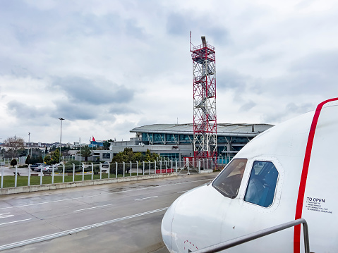 Red and white colored airport radar tower with many steps going up to the top of the antenna and the front captain's cabin of a white aircraft. Dramatic cloudy sky and air travel background, copy space.