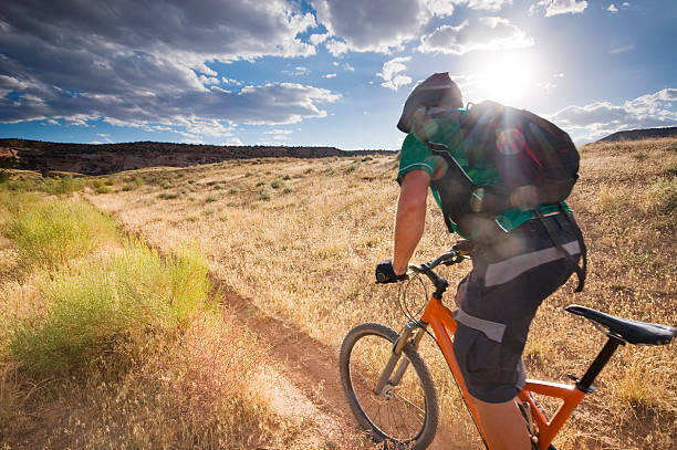 mountain biking! mountain biker speeding through the desert meadow terrain beneath a cloud filled blue sky filled with the sun during sunset turning the landscape a golden yellow.  horizontal composition with copy space. fruita colorado stock pictures, royalty-free photos & images