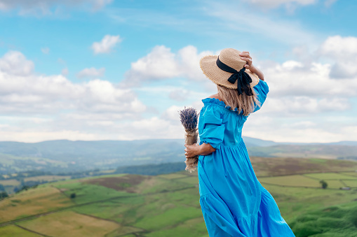 asian woman in a blue dress and a hat with a lavender bouquet in her hands on a top of mountain at summer sunset