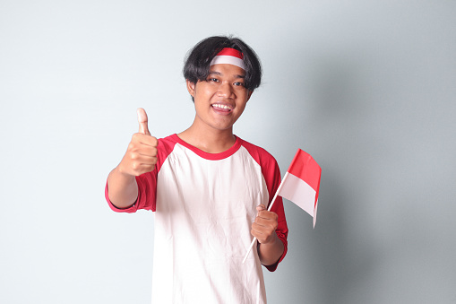 Portrait of attractive Asian man in t-shirt with red and white ribbon on head, showing product and poiting away while holding indonesian flag. Isolated image on gray background