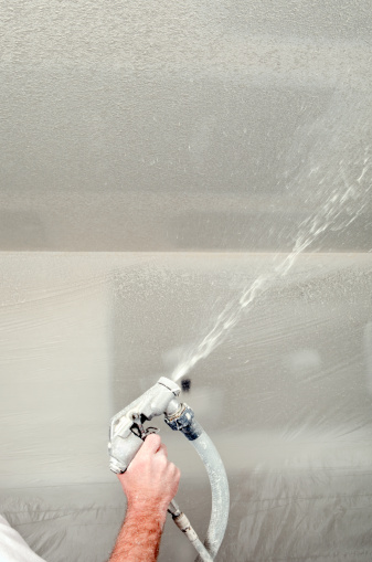 A drywall worker is spraying thick mud on the ceiling of a residential construction site. The liquid will set for a brief time and the worker will trowel over it to create a knockdown texture. The walls are covered with protective plastic and there is spray on the plastic and filling the frame.
