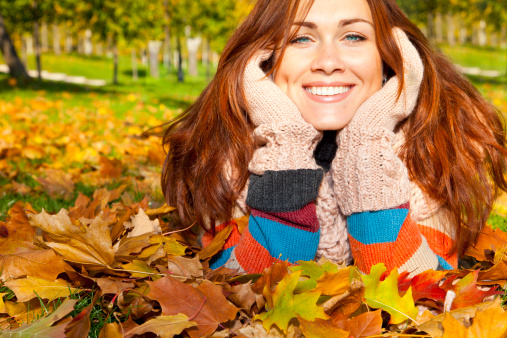 Woman with red hair laying on autumn leaves. She has gloves and a multicolored sweater.