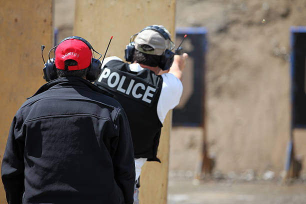 Picture of a Policeman shooting handgun in a practice field.  The police officer is wearing a police kevlar vest and has earmuffs to protect him from the sound of the handgun. The shooting target is blur and visible in the background. An insctructor is in the foreground.