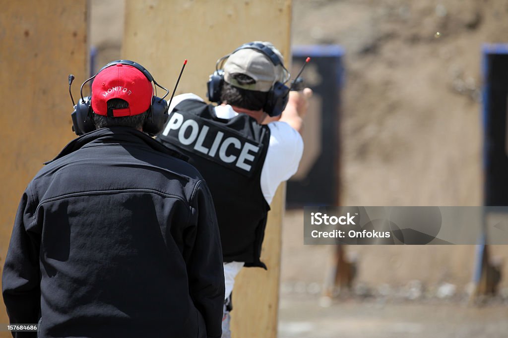 Policeman Officer Shooting 9mm Handgun with Instructor Picture of a Policeman shooting handgun in a practice field.  The police officer is wearing a police kevlar vest and has earmuffs to protect him from the sound of the handgun. The shooting target is blur and visible in the background. An insctructor is in the foreground. Target Shooting Stock Photo