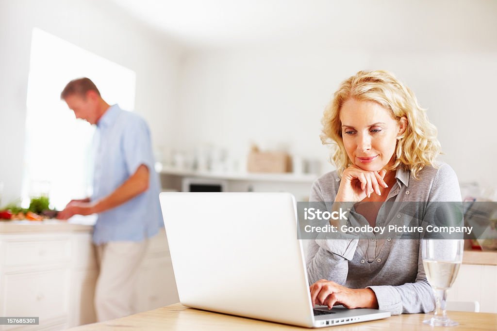Mature woman using laptop while man cooking in kitchen Mature woman using laptop while man cooking in the background Contrasts Stock Photo