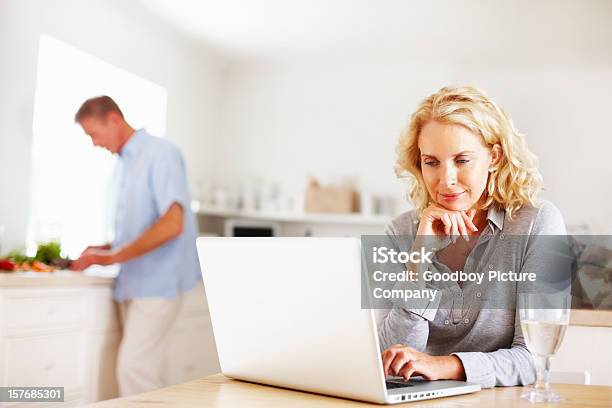 Mature Woman Using Laptop While Man Cooking In Kitchen Stock Photo - Download Image Now