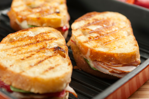 Hot panini sandwiches in the pan made with crusty, hand sliced bread, smoked ham, swiss cheese, roasted red peppers, and baby spinach leaves.