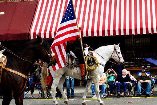 Man Holding American Flag Riding A White Horse In Small Town Parade With Spectators