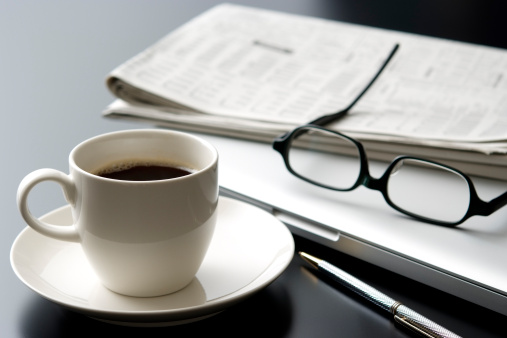 A close up of a desk at a business office.  This desk includes a cup of black coffee served in a white cup on a white saucer, a laptop computer, a pair of glasses and newspaper.