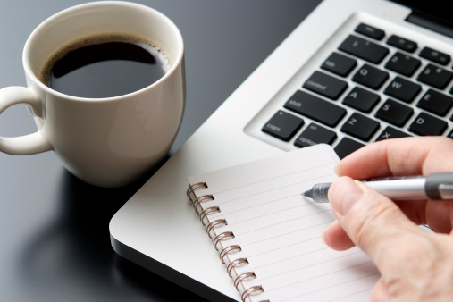 A close up of a desk at a business office.  This desk includes a cup of black coffee served in a white cup, a laptop computer.