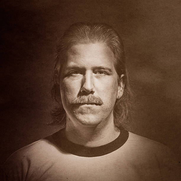 A worn and damaged photo of a serious man with slicked back hair and a bushy mustache.  Texture overlay for aged look.