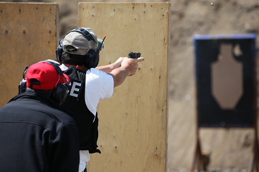 Picture of a Policeman shooting handgun in a practice field.  The police officer is wearing a police kevlar vest and has earmuffs to protect him from the sound of the handgun. The shooting target is blur and visible in the background. An insctructor is in the foreground.