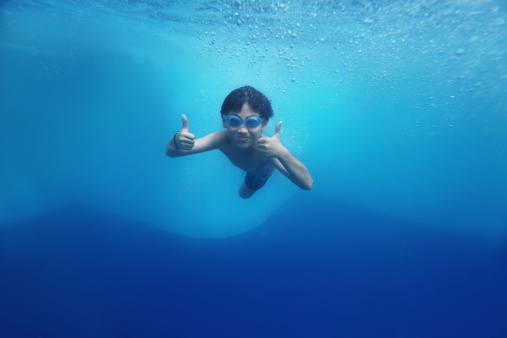 Asian boy swimming underwater moments after diving. Focus on thumbs up hands. 