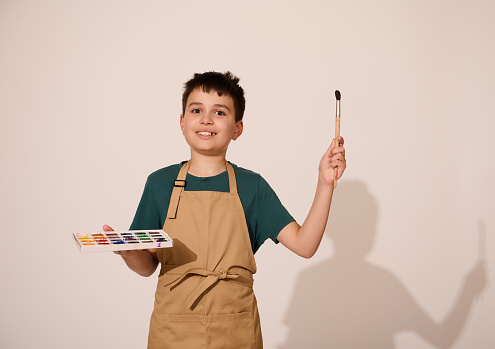 Adorable child boy enjoying art class, pointing a paintbrush on a copy space, holding a palette of colorful vibrant watercolor paints, isolated over white studio background. Creative hobby and leisure