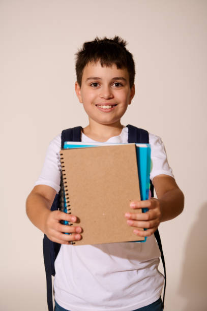 istock Smiling happy school child holding out at camera copybook with copy space for your ads., isolated over white background. 1576878280