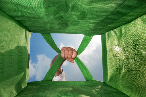 Looking up from the bottom of a green, reusable bag, Seeing a hand holding the handles. Reduce, reuse, recycle.