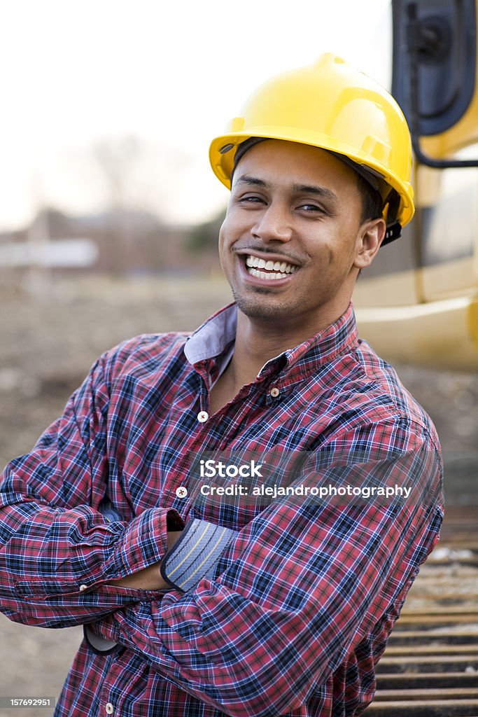 ethnic construction worker smiling A man working on a construction site with his hands crossed in front. The man is smiling and is wearing a yellow hardhat, grey and blue construction gloves, and a red and blue plaid long sleeve button up shirt. 20-29 Years Stock Photo