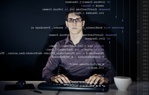 young man with glasses sitting in front of his computer, programming. the code he is working on (c#) can be seen through the screen.