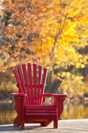 An adirondack chair in fall. Vertical image. Ontario, Canada. A lone red wooden muskoka chair on a dock. This image is taken near Algonquin Provincial Park near Deerhurst Highlands Resort. Nobody is in the image. Beautiful fall colours enhance this restful scene on a pretty lake in cottage country. Image taken with Canon 5D Mark II camera and an L series lens. 
