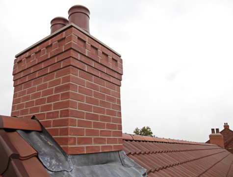 An isolated portrait of the top part a tall masonry grout red brick chimney on a black tile roof of a house during daytime with a blue sky and some clouds.