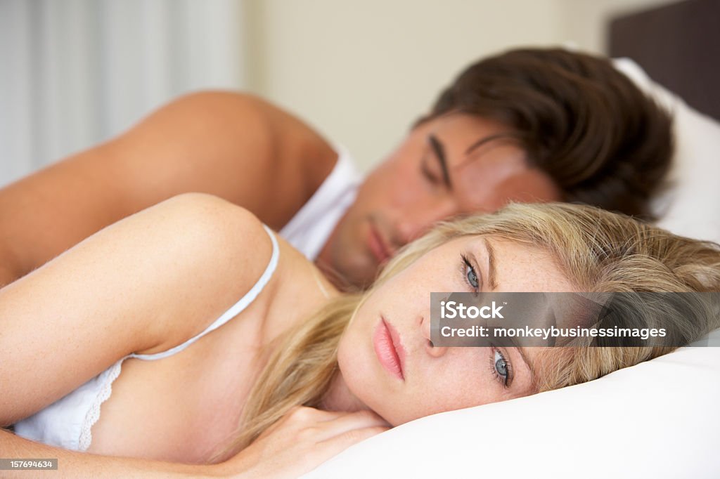 Young couple in bed, man asleep and woman looking worried Worried Young Couple Laying In Bed 20-29 Years Stock Photo