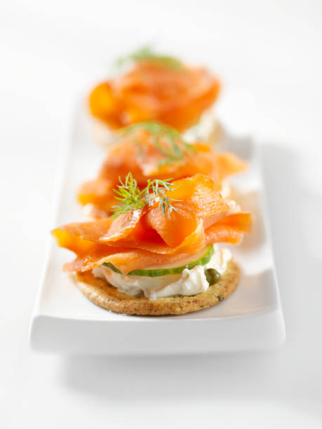 Stack of black charcoal and traditional crackers with smoked salmon, cream cheese, green salad and red currant berries on vintage metal tray over black stone background. Appetizer snack. Top view