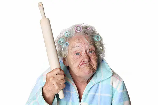 80ish senior female in rollers and plaid bathrobe is making faces and holding rolling pin like she's ready to hit you over the head. Humor image.