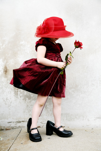 Profile of unrecognizable little girl in red velvet dress with adult oversized hat and shoes.  Dress is blowing backwards in the wind. She's holding a rose. Full body showing in stylized image that is part of an on-going series.