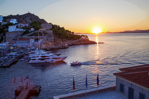 Bay of Hydra in fantastic sunset. Small boat (water taxi) from Mainland Greece approaching to harbor. Large yachts in parking lots. Idyllic vacations on Greek islands
