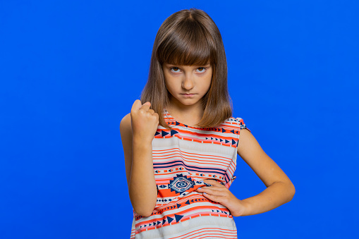 Aggressive angry mad young school girl trying to fight at camera, shaking fist, boxing with expression, punishment, abuse, rage. Preteen Caucasian bully child kid isolated on studio blue background