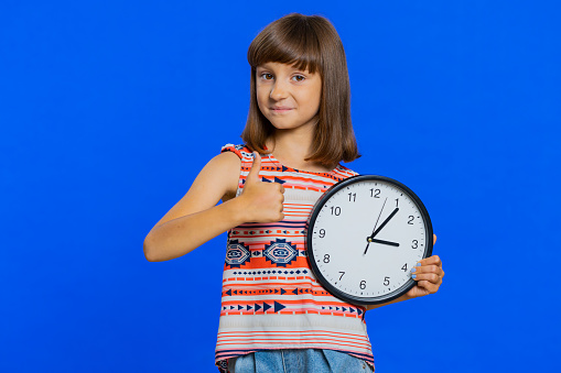 It is your time. Young school girl showing time on wall office clock, ok, thumb up, approve, pointing finger at camera, time for education study. Preteen child kid isolated on studio blue background