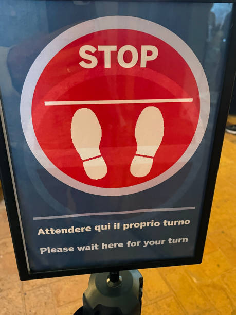 istock Close-up image of Italian 'Stop - Please wait here for your turn' vertical sign frame on belt stanchion, circular, red stop sign with footprint symbol, waiting in line, focus on foreground 1577124529