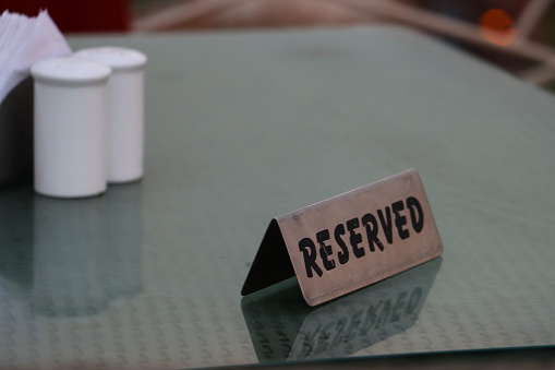 Stock photo showing close-up view of double sided tent design, brushed stainless steel reserved table sign on glass topped woven table with napkin holder of paper napkins and salt and pepper pots in restaurant.