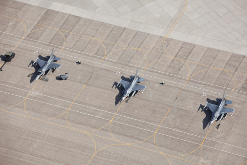 An aerial view of three F-16 fighter jets sitting on an airfield tarmac ready for flight. All have wing mounted fuel tanks and AMRAMM wingtip (air-to-air) missiles, the left two have Harm (air-to-ground) missiles. The left plane's canopy is open and the safety flags are removed for flight. The left two planes have safety flags in place. Shot from the open window of a small airplane.  http://www.banksphotos.com/LightboxBanners/Aerial.jpg http://www.banksphotos.com/LightboxBanners/Aircraft.jpg