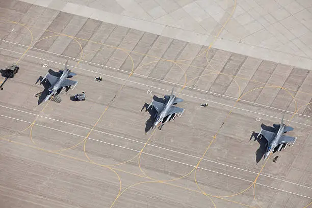 An aerial view of three F-16 fighter jets sitting on an airfield tarmac ready for flight. All have wing mounted fuel tanks and AMRAMM wingtip (air-to-air) missiles, the left two have Harm (air-to-ground) missiles. The left plane's canopy is open and the safety flags are removed for flight. The left two planes have safety flags in place. Shot from the open window of a small airplane.  http://www.banksphotos.com/LightboxBanners/Aerial.jpg http://www.banksphotos.com/LightboxBanners/Aircraft.jpg