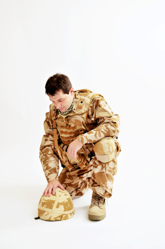 Pensive soldier, looking down and kneeling with his hand resting on his helmet. Soldier is wearing British Military desert camouflage uniform. Studio shot with a plain background.