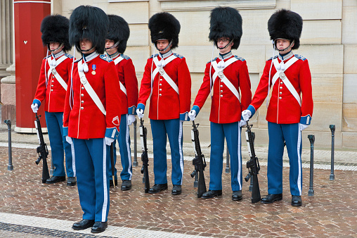 Armed soldiers guards using bearskin are marching in the front yard at Buckingham Palace, London, England. This picture was taken from outside the palace during the changing of The Queen's Guard, the infantry soldiers charged with guarding the official royal residences in the United Kingdom. A bearskin is a tall fur cap, usually worn as part of a ceremonial military uniform.